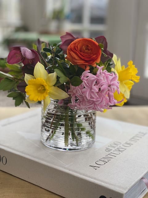 A colorful bouquet of flowers, including roses and daffodils, in a glass vase placed on top of a book on a table.