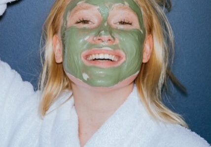 A woman in a bathrobe with a green clay mask on her face.